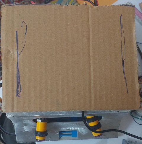 The built sumo robot: an aluminium base plate carrying two yellow cylindrical infrared proximity sensors at the front and wiring, with a low box body and a temporary cardboard top cover.
