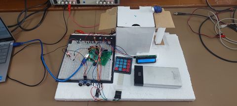 Breadboard prototype of the access box on the lab bench: ATmega8 with an AVR programmer, a 4×4 keypad, a 16×2 LCD, an HC-SR04 ultrasonic sensor, jumper wiring, and a servo-actuated cardboard enclosure standing in for the locked box.